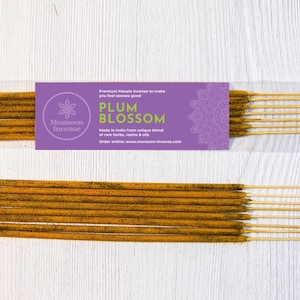 May include: Incense sticks with a purple label that reads "PLUM BLOSSOM" and "Monsoon Incense". The incense sticks are a golden brown color and are packaged in a clear plastic wrapper. The background is a white wooden surface.