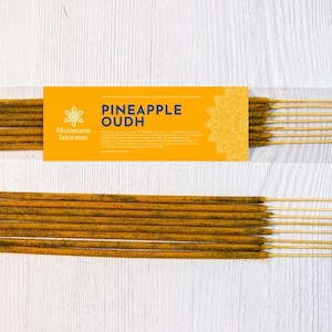 以下が含まれることがあります： Monsoon IncenseのPineapple Oudhとラベルが貼られた茶色のインセンススティックのパッケージ。花柄のロゴとテキストが記載された明るい黄色の長方形の紙帯が付いており、その下に同じ茶色のインセンススティックが並べられています。