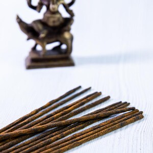 Peut inclure: Un paquet de b&acirc;tonnets d'encens bruns sur une surface en bois blanc. Une petite statue de divinit&eacute; en laiton floue se trouve en arri&egrave;re-plan.