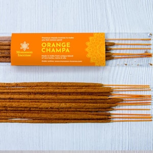 Pode incluir: Varetas de incenso Orange Champa em embalagem plástica transparente e soltas. A embalagem é laranja com o texto "Orange Champa" e "Monsoon Incense". As varetas de incenso são marrons com pontas laranjas.