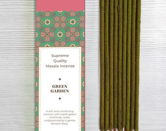 May include: A box of Bhagwan Incense with the words "Supreme Quality Masala Incense" and "Green Garden" printed on it. The box is pink and green with a floral pattern. Next to the box are several green incense sticks with red tips.