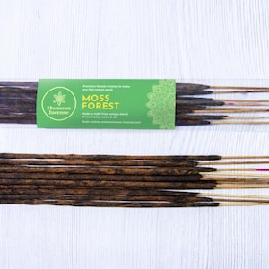 May include: Two packages of incense sticks with a green label that reads "Moss Forest" and "Monsoon Incense." The incense sticks are dark brown with red tips. The background is a white wooden surface.