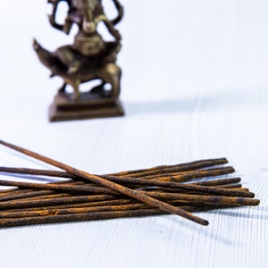 May include: A pile of brown incense sticks on a white wooden surface. A small, out-of-focus, brass statue of a deity is in the background.