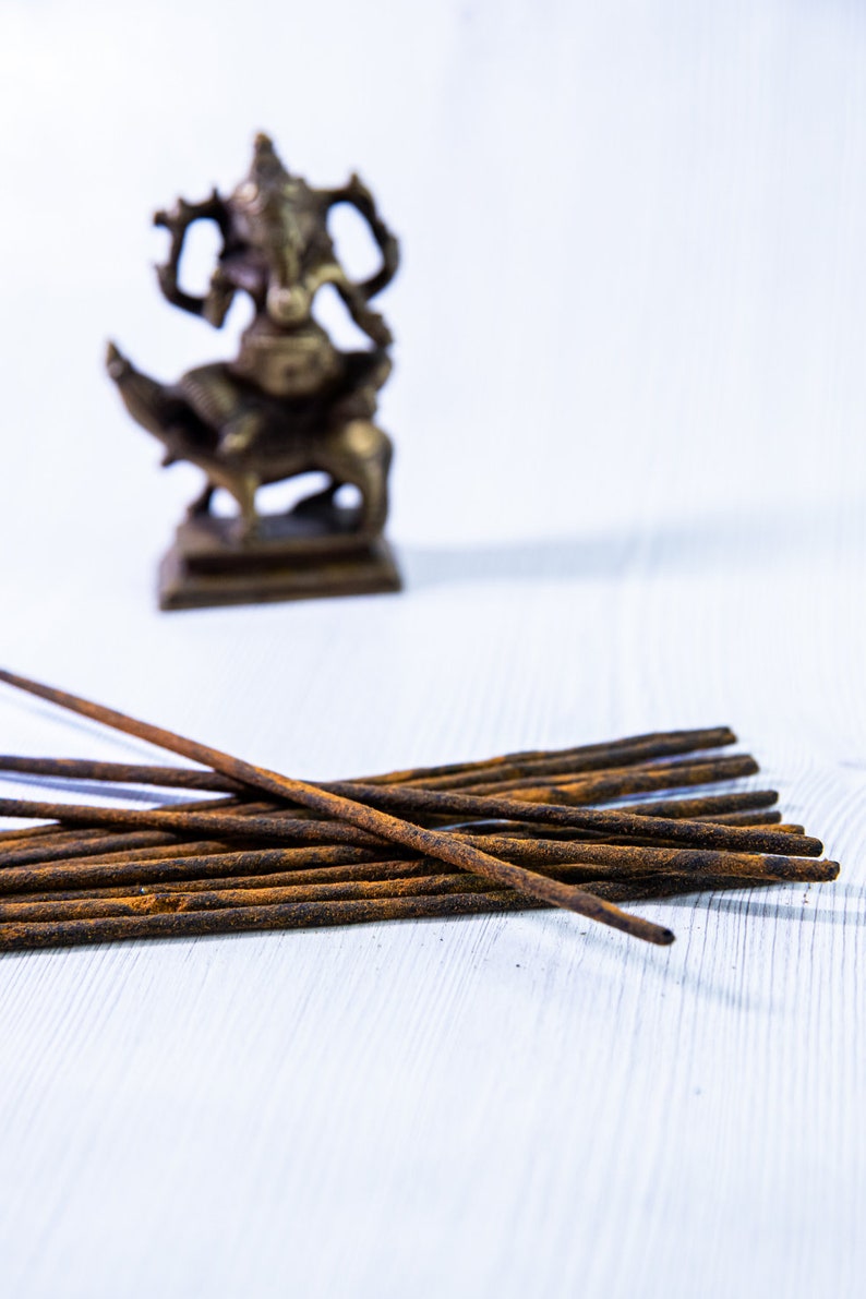 May include: A pile of brown incense sticks on a white wooden surface. A small, out-of-focus, brass statue of a deity is in the background.