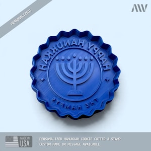 Könnte beinhalten: Ein kreisförmiger blauer Keksausstecher mit Stempel und gewelltem Rand. In der Mitte befindet sich ein erhabenes Menora-Symbol, umgeben von dem geprägten Text HAPPY HANUKKAH und THE HAMPTS. Das Design dient zum Backen personalisierter Kekse.