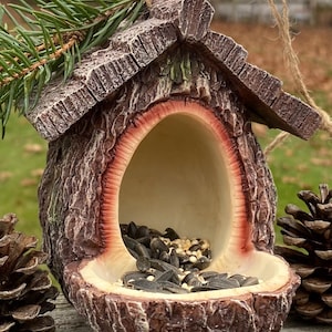 Puede incluir: Un comedero para pájaros rústico con un exterior texturizado en forma de corteza. El comedero marrón y beige está lleno de semillas para pájaros y tiene una pequeña percha. Una ramita de hoja perenne y una piña están cerca. El comedero está suspendido por una cuerda.