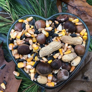 May include: A heart-shaped glass bowl filled with a variety of birdseed, including sunflower seeds, peanuts, corn kernels, and acorns.