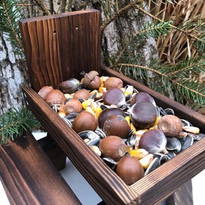 May include: A dark brown stained wooden picnic table shaped bird feeder filled with a mixture of brown acorns, chestnuts, yellow corn kernels, and black sunflower seeds. The feeder features a high backrest and two attached side benches.