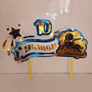 May include: A cake topper with a blue and gold banner that says "Damon" and "10". There is a gold star with a shooting star tail and a gold astronaut with the text "I need more space".
