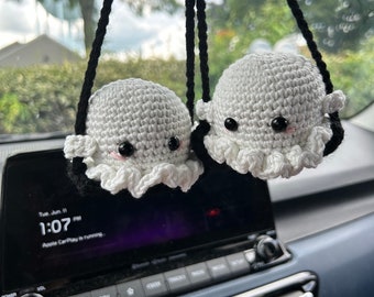 Pode incluir: Dois peluches de polvo branco de crochê pendurados em um espelho retrovisor em um carro. Os peluches têm olhos pretos e bochechas rosadas.