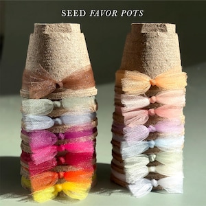 May include: Two stacks of tan fiber seed pots, each adorned with a vertical row of colorful tulle bows. The left stack features vibrant shades of brown, green, blue, magenta, pink, red, orange, and yellow. The right stack displays pastel tones.