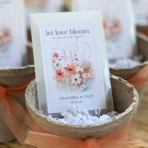 May include: Small, round, biodegradable pots filled with white paper and a packet of wildflower seeds. The packet reads "Let Love Bloom" with a floral design and the names "Chandra & Zain" and the date "08.23.25". Each pot is tied with orange tulle.