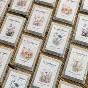 May include: A collection of wildflower seed packets with the text "let love bloom" and names with dates. The packets feature watercolor floral illustrations in various colors, including pink, orange, and blue. The packets are arranged on a wooden surface.
