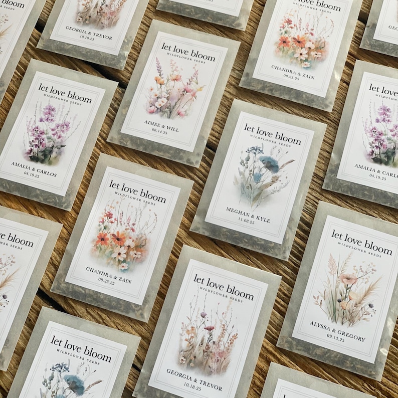 May include: A collection of wildflower seed packets with the text "let love bloom" and names with dates. The packets feature watercolor floral illustrations in various colors, including pink, orange, and blue. The packets are arranged on a wooden surface.