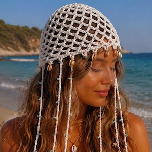 May include: A white crochet head covering with dangling strands adorned with small blue beads, seashells, and crochet circles. The headpiece is worn with a white dress, and the background shows a beach with blue water.