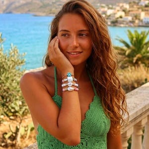 Pode incluir: Um top halter de crochê verde com um design de malha quadrada e bainha recortada, combinado com uma pulseira de contas composta por pérolas barrocas brancas, pequenas contas circulares coloridas e uma única conta azul em forma de flor.