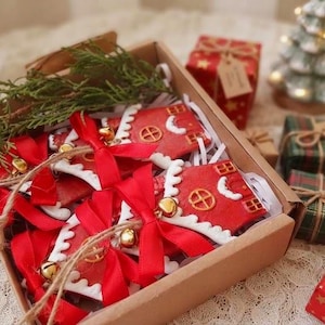 Puede incluir: Una caja de regalo de cartón que contiene varios adornos rojos con forma de casa y detalles de glaseado blanco, cada uno atado con una cinta roja y un pequeño cascabel dorado. Una rama de cedro verde está dentro de la caja, rodeada de pequeños regalos.