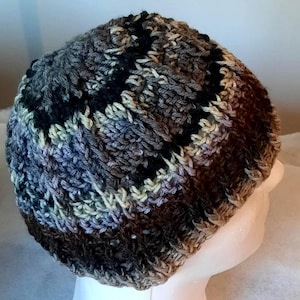 May include: A handmade, ribbed knit beanie in a mix of neutral colors. The hat features a blend of brown, black, gray, and cream yarns, creating a textured, multi-toned appearance. The beanie is designed to fit snugly and provide warmth.
