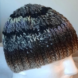 May include: A hand-knitted beanie hat in a mix of colors including black, gray, and brown. The hat has a ribbed texture and a gradient effect, transitioning from dark to light shades. The yarn appears to be thick and warm, suitable for cold weather.