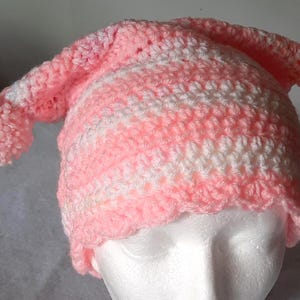 May include: A handmade crochet hat in pink and white. The hat features a long, pointed shape with two pom-poms at the ends. The hat is made with a soft, textured yarn and is likely designed for warmth and style.