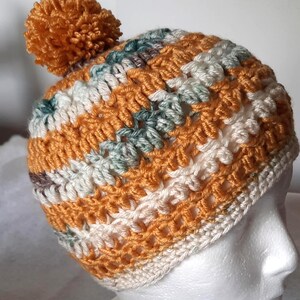 May include: A handmade crochet beanie in shades of orange, cream, and teal, topped with a matching orange pom-pom. The hat features a textured stitch pattern, perfect for cold weather. This cozy winter hat is a stylish accessory.