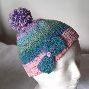 May include: A handmade crochet hat in a rainbow of colors, including pink, teal, and purple. The hat features a large pom-pom on top and a teal bow with a small purple flower accent. The hat is designed to fit a child's head.