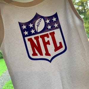 Op de afbeelding: Witte babyromper met een groot NFL-logo in rood, blauw en wit. Het logo toont een voetbal, sterren en de letters "NFL". De romper heeft een crèmekleurige rand rond de hals en de armsgaten.
