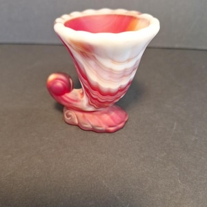 May include: A decorative glass cornucopia vase in shades of red, white, and orange. The vase has a flared, ruffled rim and a curled, horn-like design. The base is a matching color and shape.