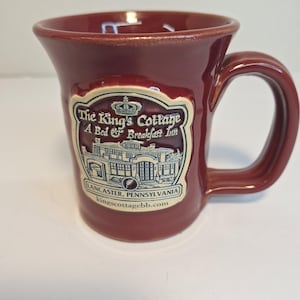 May include: A maroon ceramic mug with a curved handle. The mug features a white and blue label with the text "The King's Cottage A Bed & Breakfast Inn" and the address "Lancaster, Pennsylvania" and the website address.