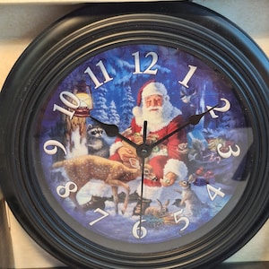May include: Round black clock with a winter scene featuring Santa Claus surrounded by woodland animals. The clock face has white numerals and black hands. The brand name "Reflective Art Inc." is visible at the top.