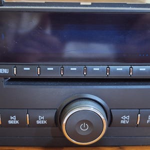 May include: A black car stereo with a digital display, buttons for FAV, MENU, BAND, SEEK, EQ, CAT, and CD/AUX. It has a large central knob with a power symbol and a smaller knob with a musical note symbol.
