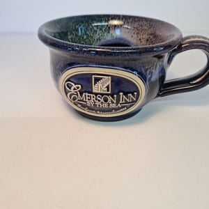 May include: A ceramic mug with a dark blue exterior and a glossy finish. The mug features a handle and an oval emblem with the text "Emerson Inn by the Sea, Rockport, Massachusetts". The interior has a blend of blue, green, and brown hues.