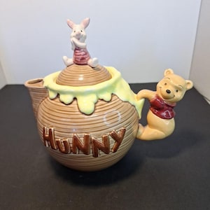 May include: A Winnie the Pooh themed teapot. The teapot is brown with the word "Hunny" in brown letters. Piglet sits on the lid, and Winnie the Pooh is the handle. The teapot has a yellow honey-like design around the lid.