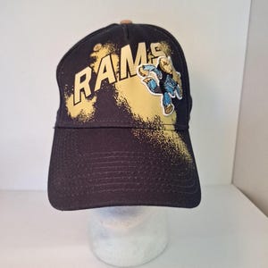 May include: Black baseball cap with the word "RAMS" in gold lettering and a gold splatter design. A cartoon figure in blue and gold is on the right side of the cap.