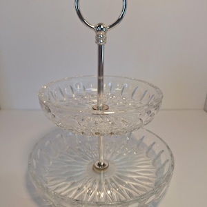 May include: A two-tiered serving tray with a silver handle and stem. The clear glass plates have a starburst pattern. The top tier is smaller than the bottom tier. The handle is a silver ring.