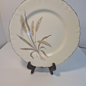 May include: A white ceramic plate with a scalloped edge and a silver rim. The plate features a wheat stalk design in shades of brown and gold. The plate is displayed on a dark brown stand.