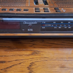 May include: A vintage Chronomatic 251 alarm clock with a wood-grain finish. The clock features an AM/FM radio dial, a digital display, and control buttons. The face of the clock displays the text "Chronomatic 251".