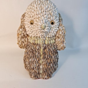 May include: A handcrafted sculpture of a creature, meticulously crafted from various seashells. The head and ears are composed of small, white shells, while the body features a textured pattern of brown and tan shells. The eyes are golden, and the nose is a small, light-colored shell.