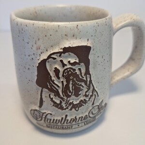 May include: Cream-colored ceramic mug with a brown dog illustration and the text "Hawthorne Inn Restaurant Lodge." The mug has a speckled finish and a curved handle. A great gift for dog lovers.