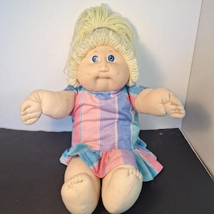 May include: A soft-sculpture doll with light skin, bright blue eyes, and dimpled cheeks. The doll has blonde yarn hair styled in a high ponytail and wears a short-sleeved dress featuring vertical stripes in shades of pink, purple, and light blue.