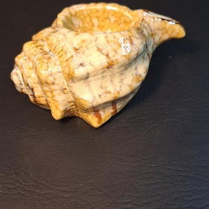 May include: A ceramic seashell with a light brown and white marbled glaze. The seashell has a deep, rounded interior and a pointed tip.