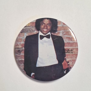 May include: Round button featuring a portrait of Michael Jackson in a black tuxedo with a bow tie. The background shows a brick wall. The button is a collectible item.