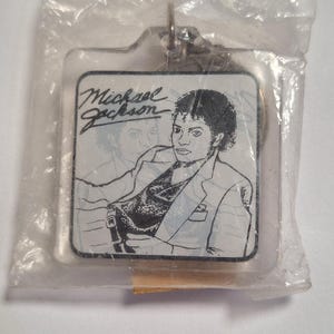 May include: A square keychain featuring a black and white image of Michael Jackson, with his name in cursive. The keychain is encased in a clear plastic wrapper, with a metal ring for attachment.