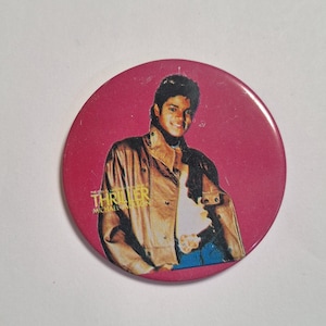 May include: A round, pink button featuring a portrait of Michael Jackson in a brown jacket. The button has the word "Thriller" and "Michael Jackson" printed on it.