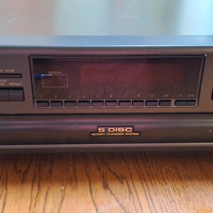 Puede incluir: Un cambiador de discos compactos Technics SL-PD607 gris oscuro. El panel frontal presenta una pantalla, botones y el texto "5 DISC COMPACT DISC CHANGER SYSTEM". La unidad se apoya en cuatro patas pequeñas.