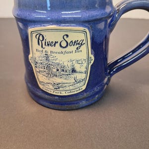 May include: A blue ceramic mug with a handle. The mug features a rectangular label with the words "River Song Bed & Breakfast Inn" and a black and white illustration of a building and trees. The text "Estes Park, Colorado" is also on the label.