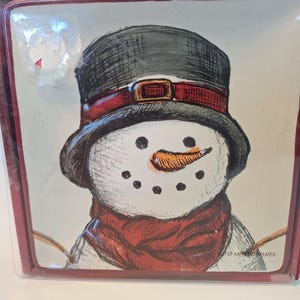 May include: Square appetizer plate featuring a snowman illustration. The snowman has a black top hat, a red scarf, and an orange carrot nose. The plate is framed with a red border. Text on the plate reads "SET OF 4 APPETIZER PLATES."