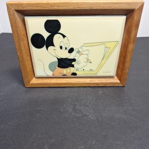 May include: A framed ceramic tile featuring Mickey Mouse drawing. The artwork shows Mickey in black, white, and tan, holding a pencil and sketching on a notepad. The frame is made of light brown wood, and the tile has a cream-colored background.