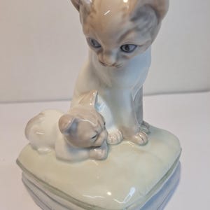 May include: A porcelain figurine of two cats. One cat is sitting upright, and the other is lying down on a pillow. The cats are light gray and white with blue eyes. The pillow is light yellow and blue.