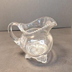 May include: Clear glass creamer with a round body, a handle, and a ruffled rim. The creamer has a decorative pattern on the base and sits on three small feet. The glass is transparent, allowing light to pass through.
